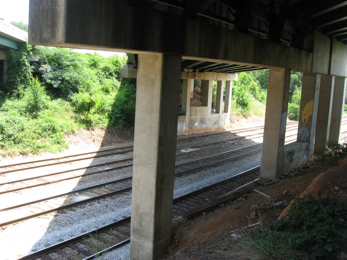 A view from beneath a concrete bridge, showcasing two sets of railway tracks running parallel to each other. The area is surrounded by dense greenery and underbrush, with some graffiti visible on the bridge's supporting columns. Sunlight casts shadows across the tracks, creating a contrast between light and shade. Chase Street mountain bike trail.