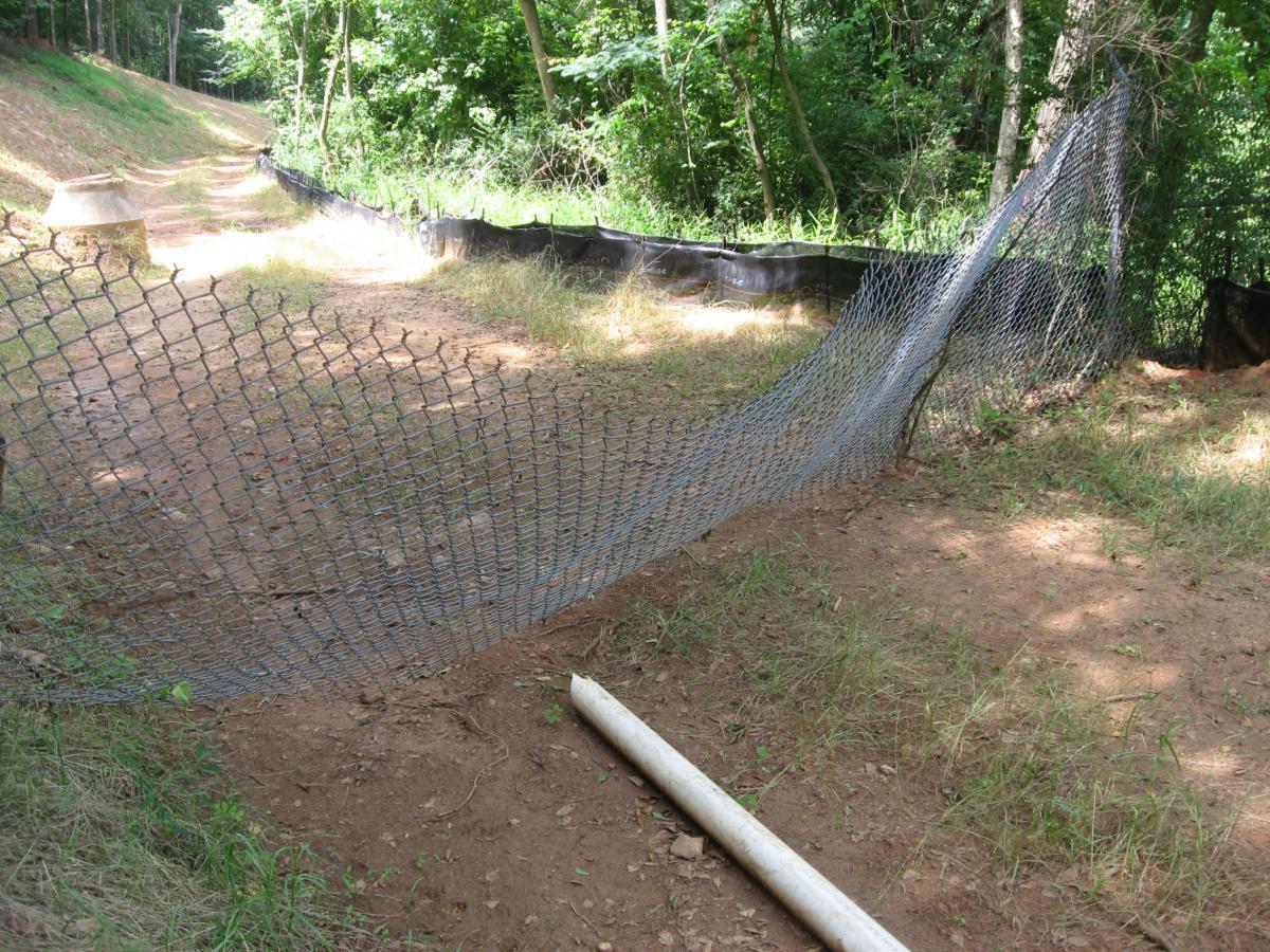 A partially damaged wire fence along a dirt pathway, with green grass and trees in the background. A white pipe lies on the ground near the fence. Oconee River Greenway mountain bike trail.