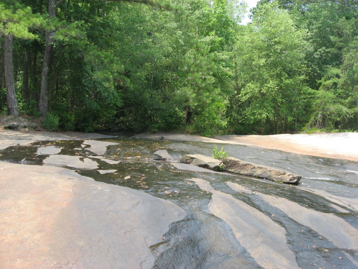 A scenic view of a rocky landscape featuring a small stream of water flowing over smooth, weathered rocks. Lush green trees and foliage border the area, creating a natural, tranquil setting. Tribble Mill Park mountain bike trail.