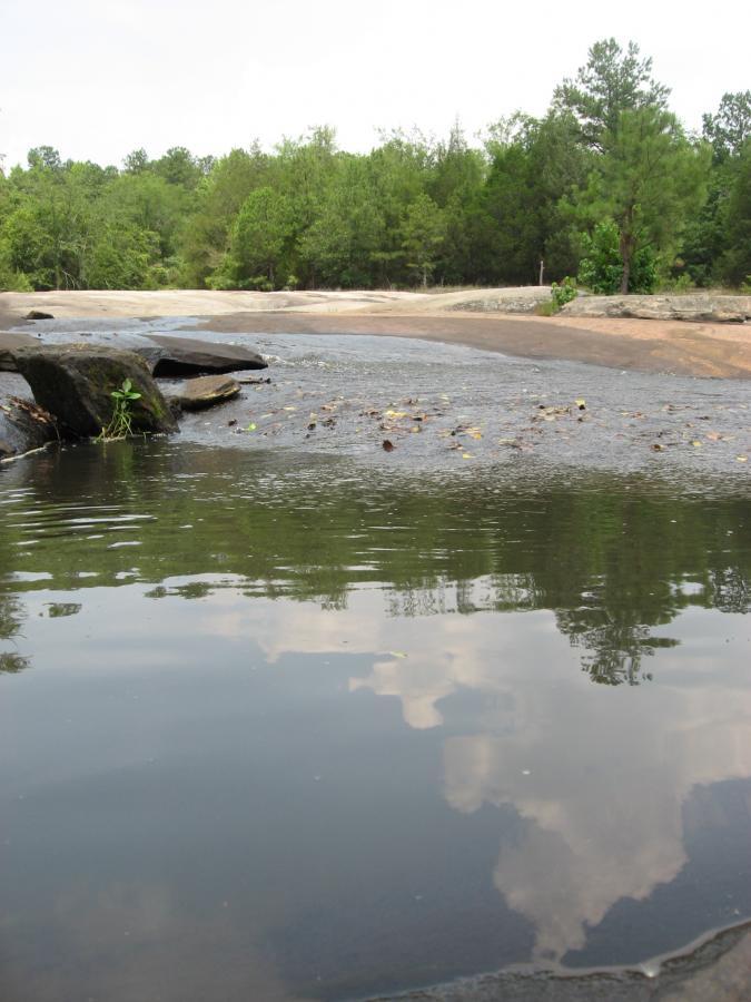 A tranquil outdoor scene featuring a calm river with reflections of clouds and trees in the water. Smooth, flat rocks are partially submerged, surrounded by greenery on the banks. The sky is overcast, creating a serene atmosphere in this natural setting. Tribble Mill Park mountain bike trail.