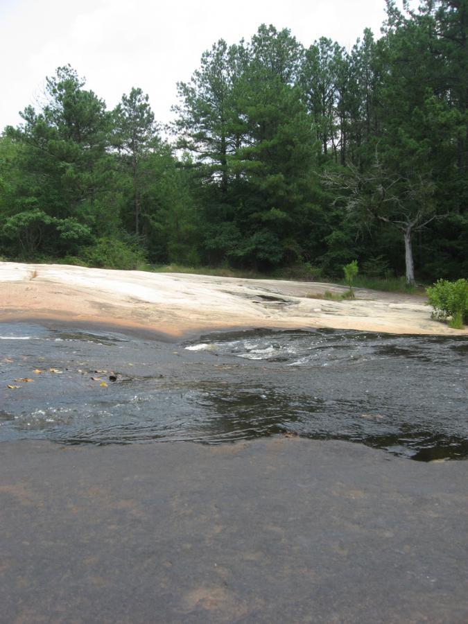A view of a rocky terrain with a flowing stream of water in the foreground, surrounded by lush green trees. The landscape features a large flat rock surface that slopes gently towards the water, creating a peaceful natural setting. Tribble Mill Park mountain bike trail.