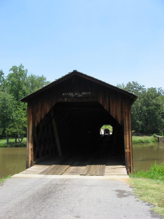 A wooden covered bridge, labeled "Watson Mill Bridge est. 1885," spans over a peaceful body of water. The entrance of the bridge is visible, showcasing the interior structure and the sunlight creating a soft glow. Surrounding the bridge are lush green trees and well-maintained grass, under a clear blue sky. Watson Mill Bridge State Park mountain bike trail.