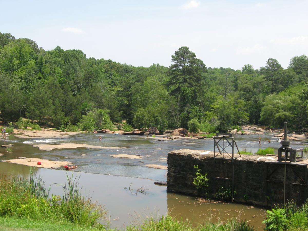 A serene landscape featuring a river surrounded by lush greenery. People can be seen enjoying the water on rocky banks under a clear blue sky. A structure made of stone and metal is situated on the right side, adding to the natural scenery. Watson Mill Bridge State Park mountain bike trail.