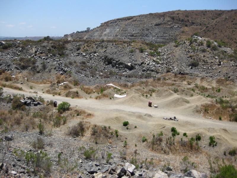 A landscape view of a barren, rocky area featuring dirt paths and sparse vegetation. The foreground includes small dirt mounds and a couple of barrels, while hilly terrain rises in the background under a clear blue sky. Mission Trails mountain bike trail.