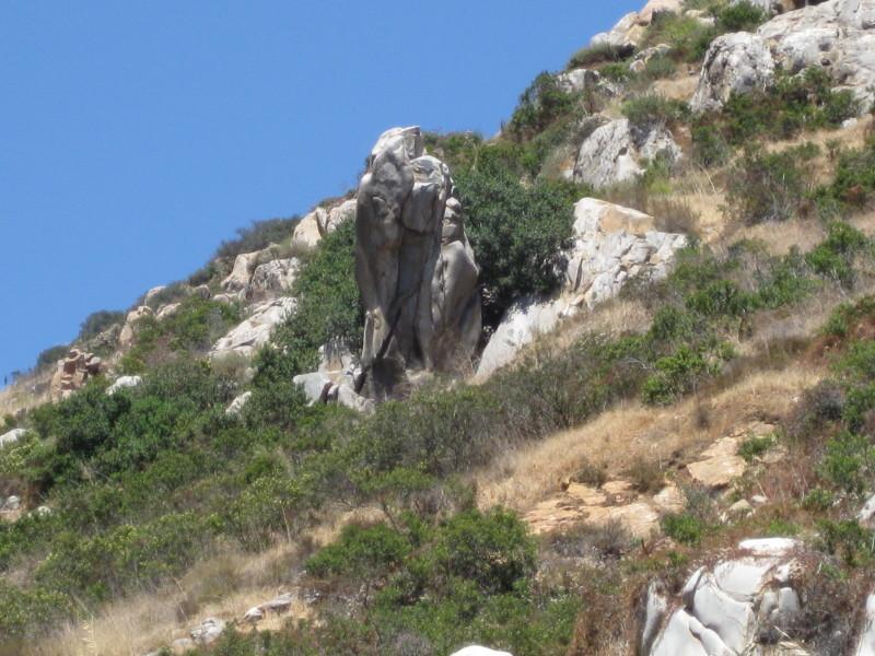 Rocky hillside with a prominent stone formation surrounded by greenery and dry grass under a clear blue sky. Mission Trails mountain bike trail.