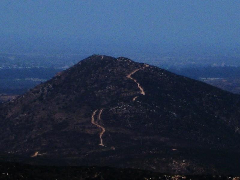A distant view of a mountain with a winding trail leading to the summit, set against a hazy blue sky. The landscape features rocky terrain and sparse vegetation. Mission Trails mountain bike trail.
