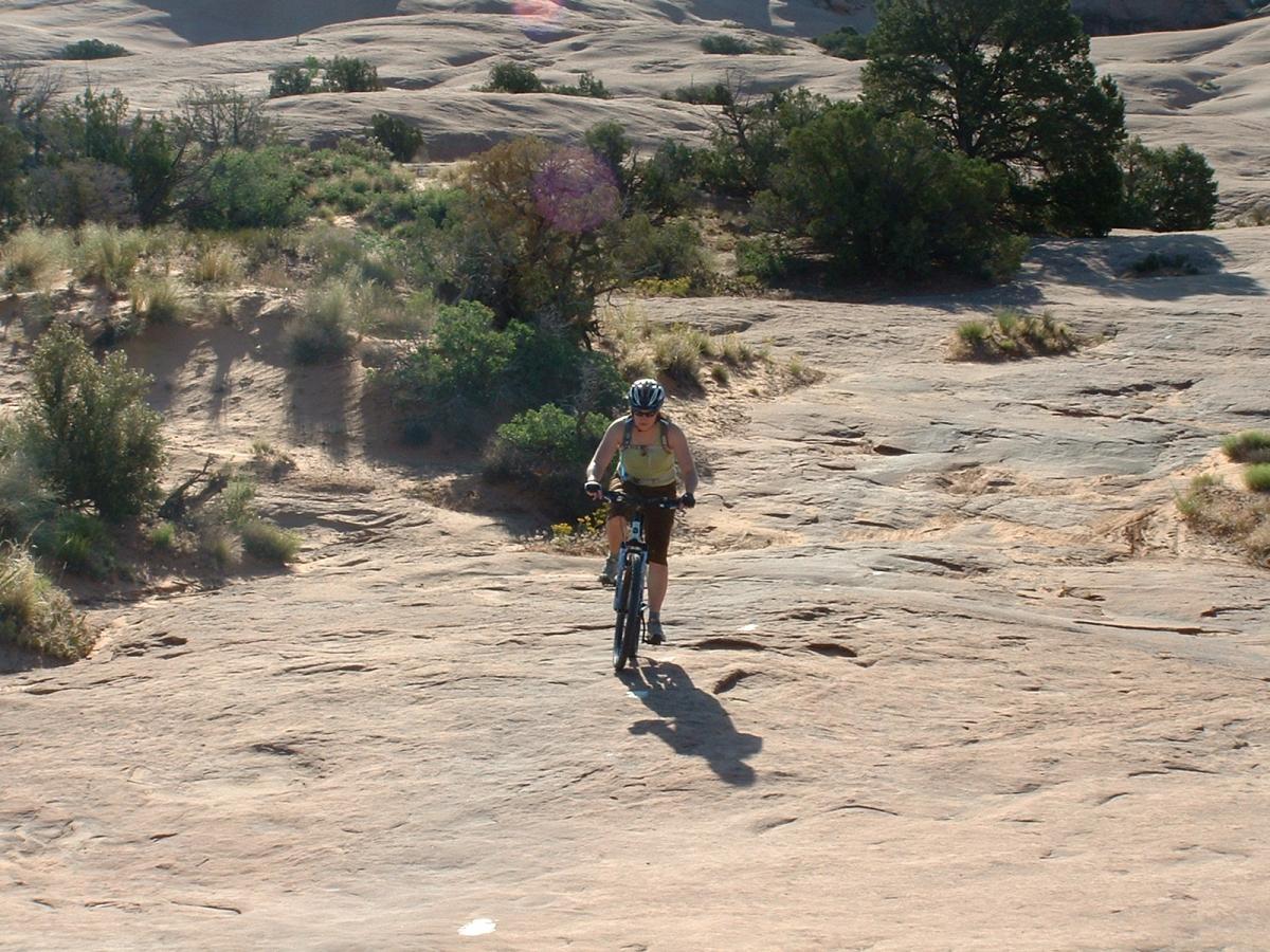 A cyclist riding a mountain bike on a rocky trail surrounded by sparse vegetation and rugged terrain. The sun is shining, and the landscape features a mix of sandy ground and rocky outcrops. Slickrock mountain bike trail.