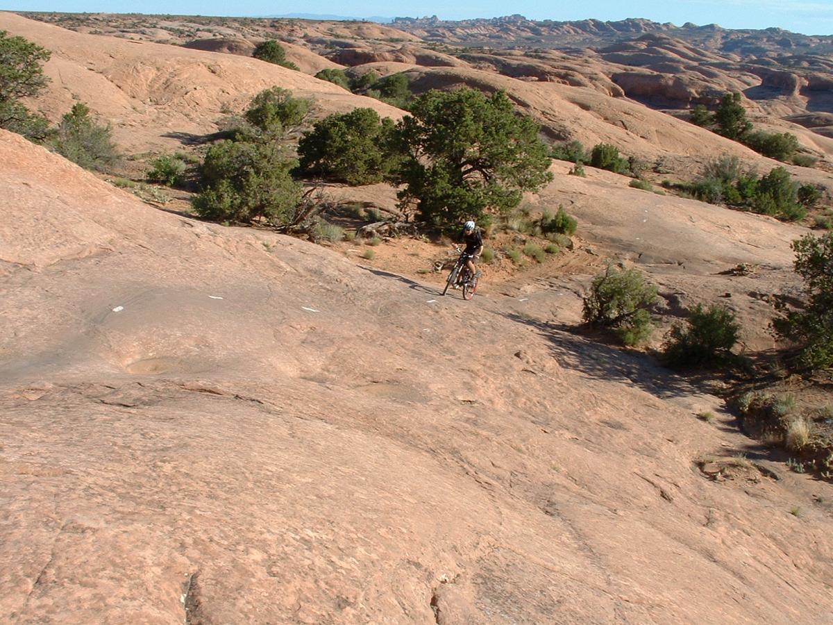 A mountain biker navigating rocky terrain on a rugged landscape, featuring sparse vegetation and rolling hills in the background. Slickrock mountain bike trail.