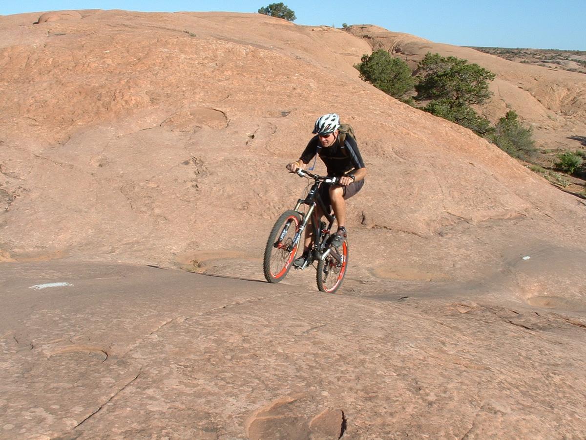 A cyclist navigating a rocky terrain on a mountain bike, showcasing outdoor biking skills on a sunlit day. The landscape features large, smooth boulders and sparse vegetation in the background. Slickrock mountain bike trail.