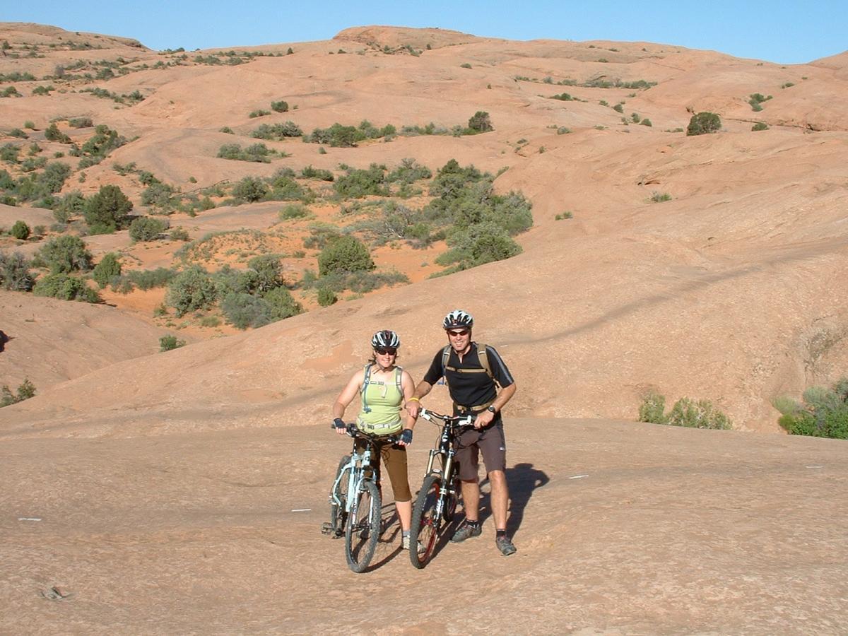 A man and a woman stand beside their mountain bikes on a rocky terrain with rolling hills and sparse vegetation in the background, under a clear blue sky. Slickrock mountain bike trail.