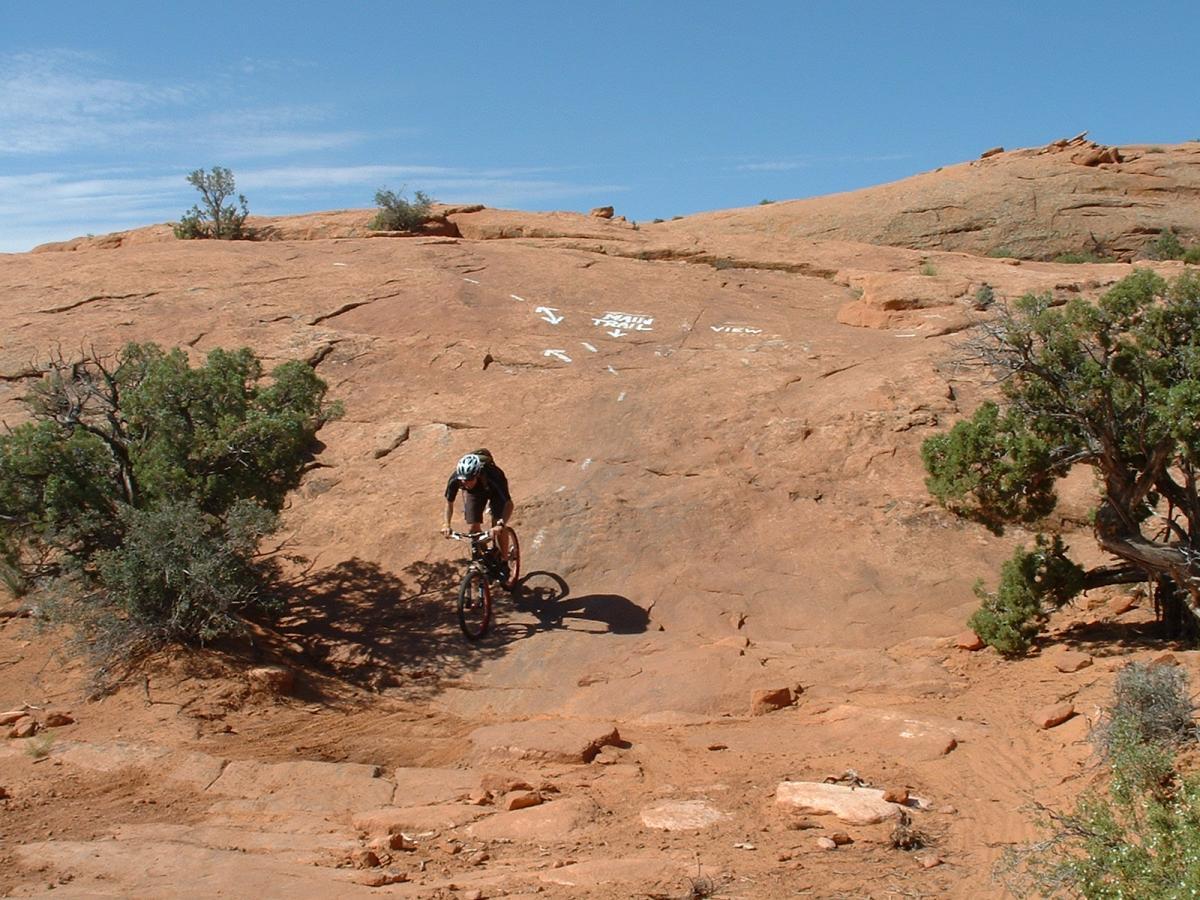 A mountain biker navigates a rocky terrain under a clear blue sky, surrounded by sparse vegetation and red rock formations. The ground is rugged and uneven, indicating a challenging biking route. White arrows are visible on the rocks, possibly marking the biking trail. Slickrock mountain bike trail.