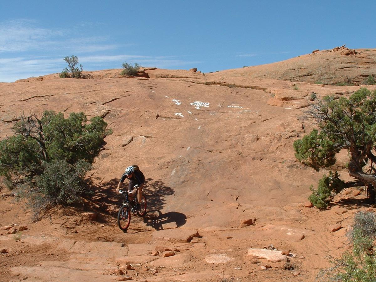 A mountain biker navigating a rocky terrain with red sandstone under a clear blue sky. Sparse vegetation, including small bushes, can be seen alongside the trail marked with directional arrows. Slickrock mountain bike trail.