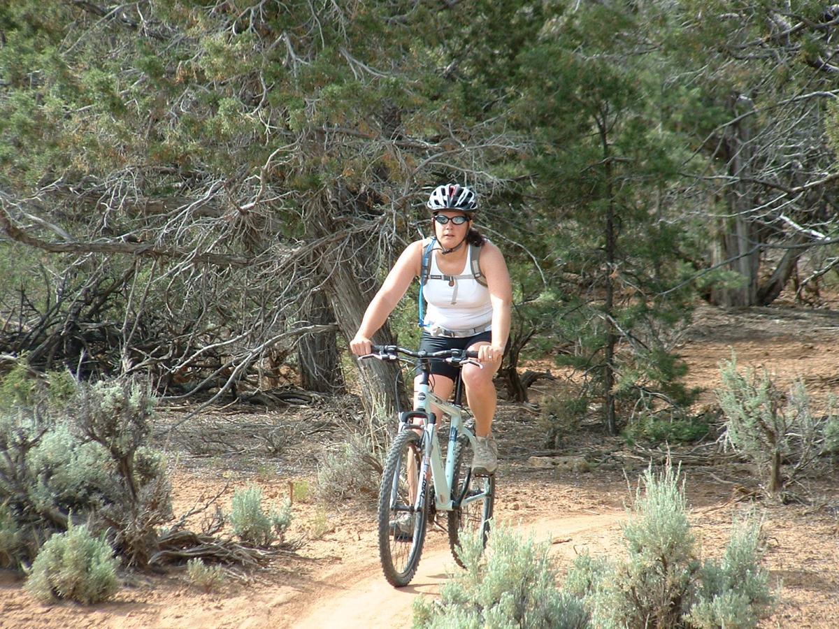 A person riding a mountain bike on a sandy trail surrounded by trees and shrubs. They are wearing a helmet and sunglasses, dressed in a tank top and shorts, and are focused on the path ahead. Phil's World mountain bike trail.