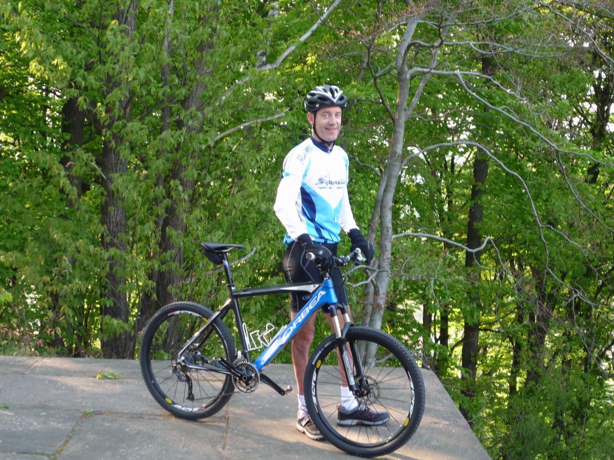 A cyclist wearing a blue and white jersey and a black helmet stands next to a blue mountain bike on a paved surface, surrounded by lush green trees in a sunny outdoor setting. Tower Trail mountain bike trail.