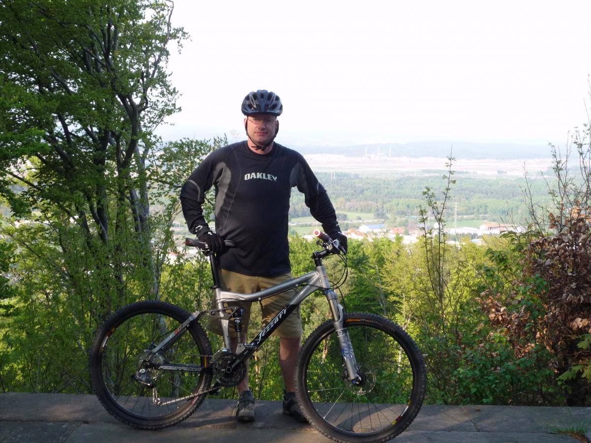 A person in athletic clothing stands beside a mountain bike, posing for the camera in a scenic outdoor setting with trees and a distant landscape. The individual is wearing a black shirt with "Oakley" printed on it and a helmet. The background showcases greenery and a valley view. Tower Trail mountain bike trail.