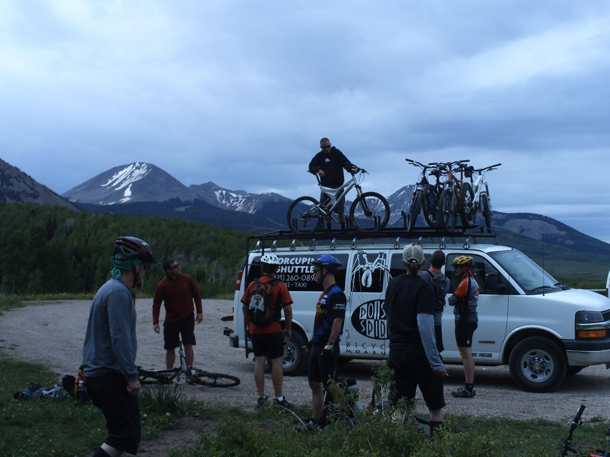 A group of mountain bikers gathered near a white shuttle van parked on a gravel road, with several bicycles on the roof. In the background, there are green trees and mountains, some capped with snow under a cloudy sky. The bikers, wearing helmets and riding gear, appear to be preparing for a ride. Porcupine Rim mountain bike trail.