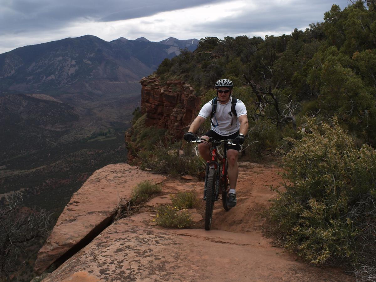 A mountain biker riding along a rocky trail with a scenic view of mountains in the background. The sky is cloudy, and the terrain features green shrubs and exposed rock surfaces. Porcupine Rim mountain bike trail.