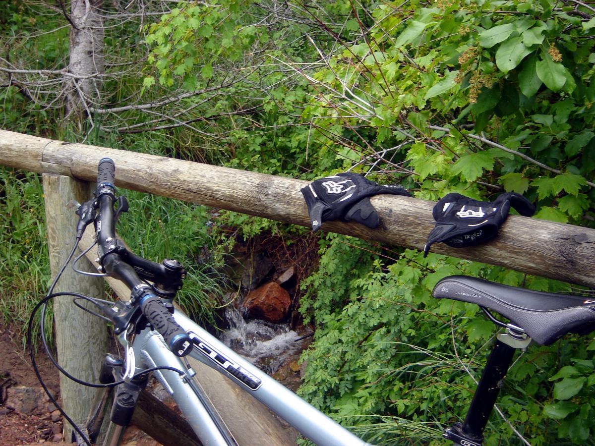 A mountain bike resting against a wooden railing near a stream, with cycling gloves placed on the railing. Lush green foliage surrounds the scene. Chimney Gulch mountain bike trail.