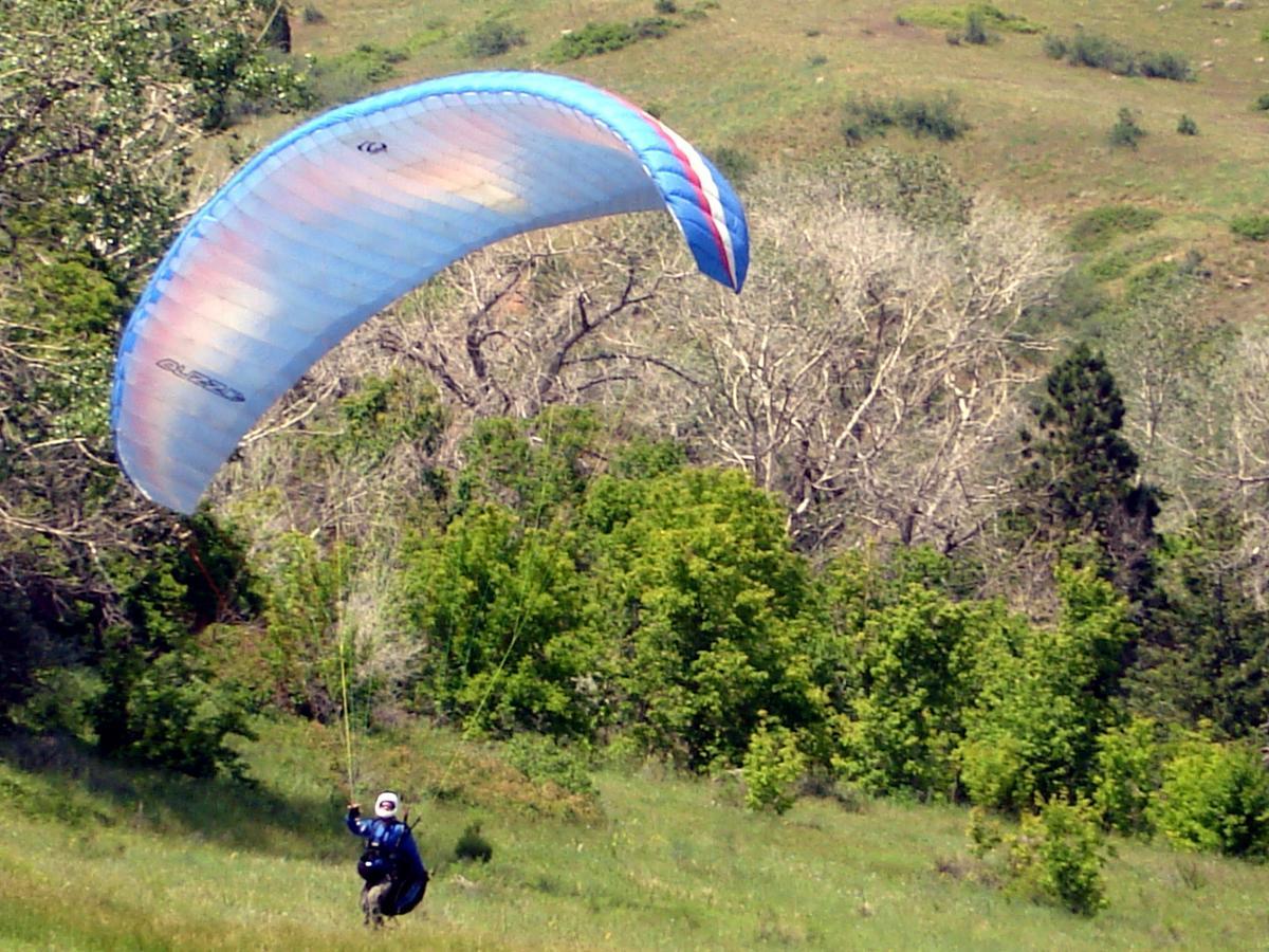 A person dressed in a blue jacket and helmet is paragliding, holding onto the controls of a colorful, blue and red paraglider. The scene is set in a lush, green landscape with trees in the background and a clear sky above. Chimney Gulch mountain bike trail.