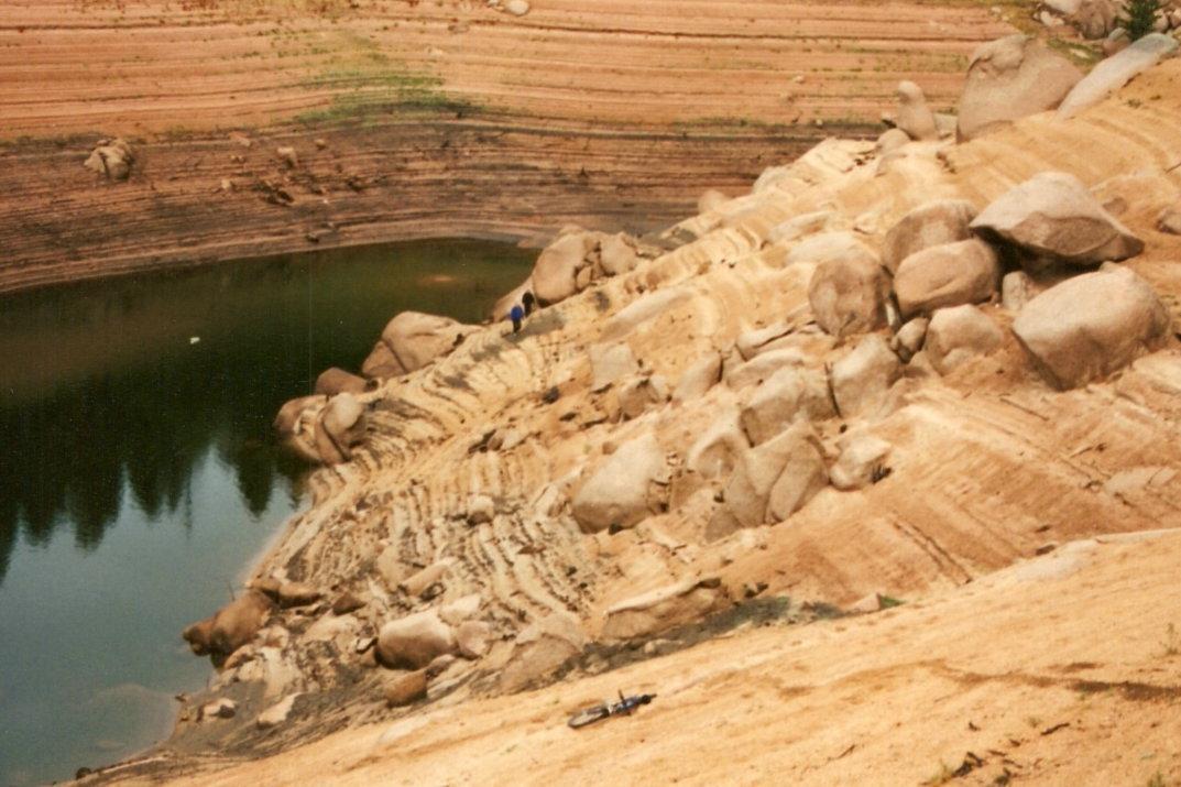 A barren landscape featuring a rocky sloped area leading down to a small body of water. The terrain is characterized by layered, sandy sections with large boulders scattered throughout. In the distance, two people are visible, exploring the rocky bank near the water. Rampart Reservoir mountain bike trail.
