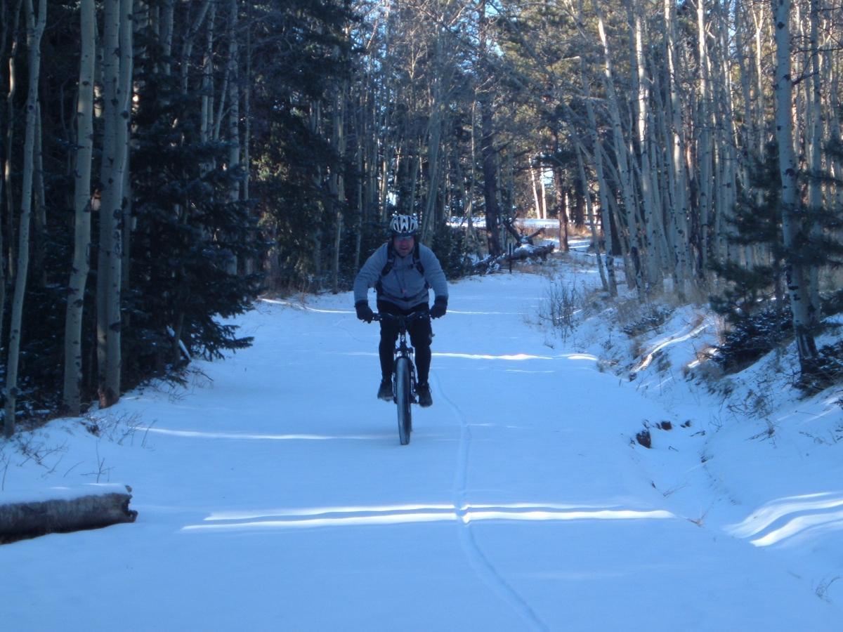 A person riding a mountain bike along a snow-covered trail surrounded by tall trees in a winter forest. The cyclist is wearing a helmet and a jacket, and the path shows tracks indicating recent activity in the snow. Rampart Reservoir mountain bike trail.