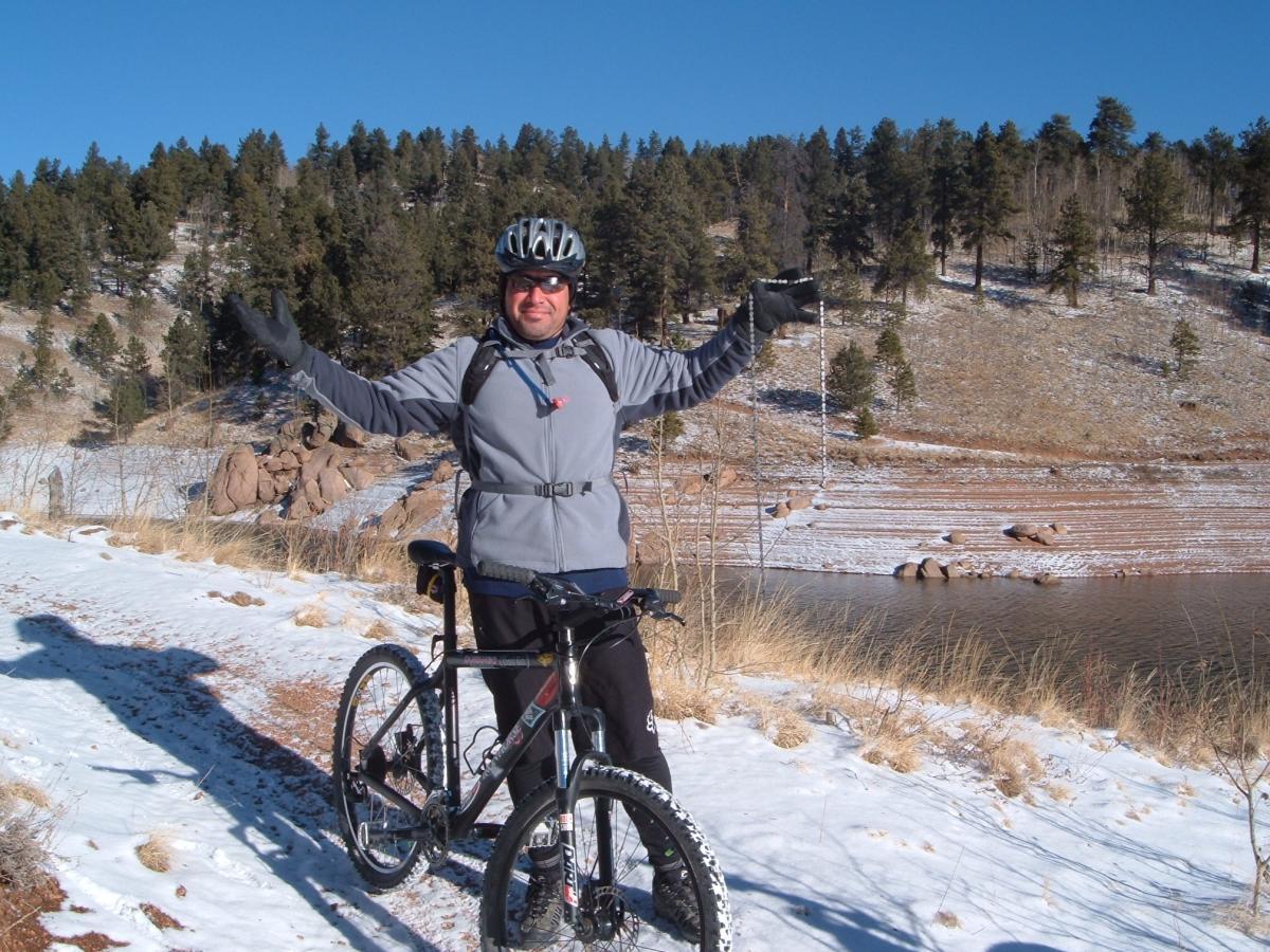 A person in a helmet and cycling gear stands next to a mountain bike on a snow-covered path, with their arms raised in a joyful gesture. In the background, there are trees and rocky terrain, with a body of water visible nearby under a clear blue sky. Rampart Reservoir mountain bike trail.