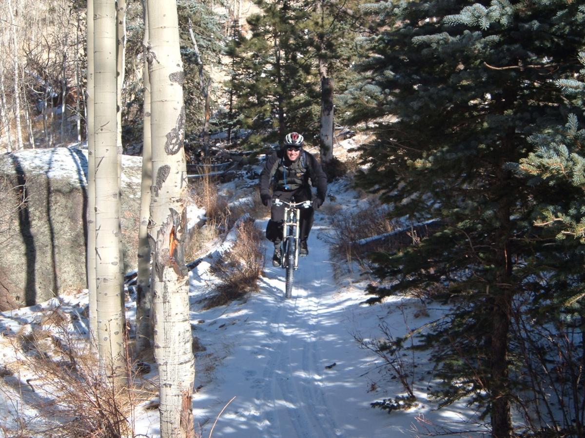 A cyclist riding a mountain bike on a snowy trail surrounded by tall trees in a forested area. Rampart Reservoir mountain bike trail.