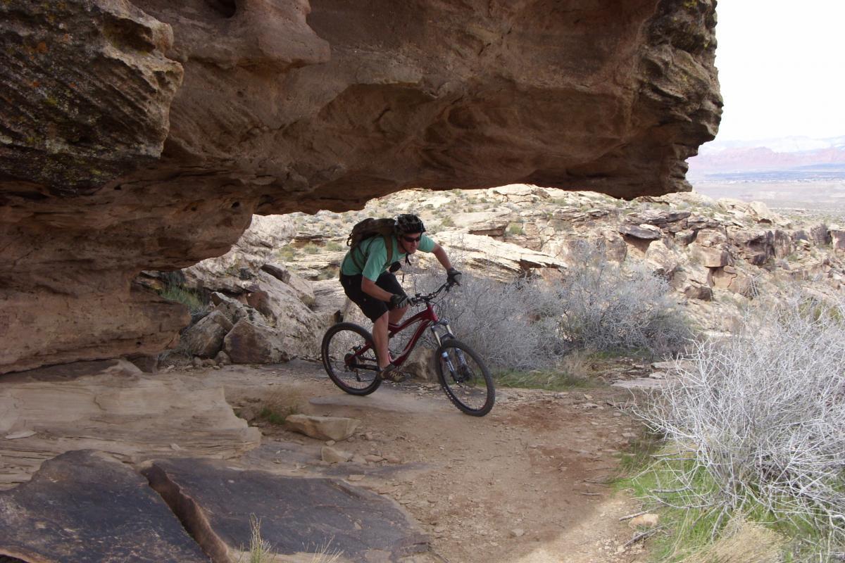 A mountain biker navigates a rocky trail beneath a large overhang in a rugged landscape, surrounded by sparse vegetation and distant hills. Zen Trail mountain bike trail.