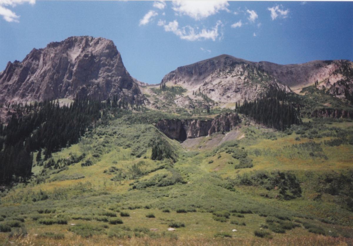 A scenic view of rugged mountains under a bright blue sky, featuring lush green hills and patches of trees in the foreground. The landscape showcases a diverse terrain with rocky peaks and varying vegetation. Trail 401 mountain bike trail.