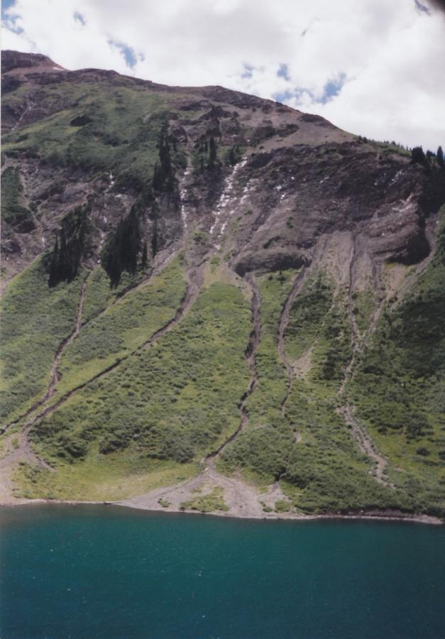 A scenic view of a lush green hillside with visible erosion patterns leading down to a tranquil blue body of water. The sky is partly cloudy, enhancing the natural beauty of the landscape. Trail 401 mountain bike trail.