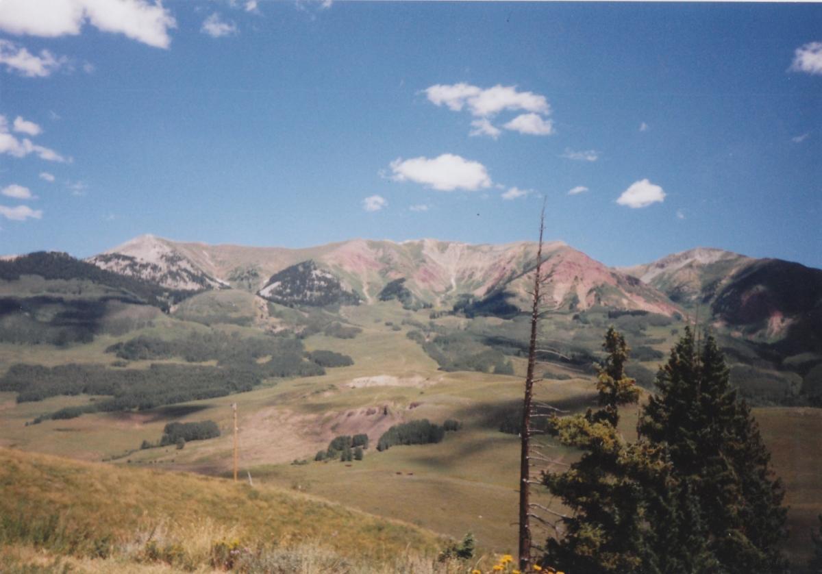 A scenic view of colorful mountains under a clear blue sky, featuring a mix of green forests and grassy valleys in the foreground. Some clouds dot the sky, and a few trees are visible in the lower right corner, adding to the natural landscape. Trail 401 mountain bike trail.