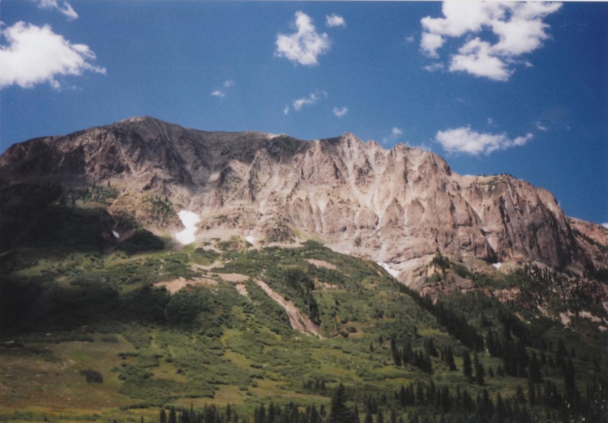 A picturesque view of a rugged mountain peak surrounded by lush green vegetation and trees, under a clear blue sky with scattered white clouds. The sunlight highlights the texture of the rocky slopes and patches of snow in the shaded areas. Trail 401 mountain bike trail.
