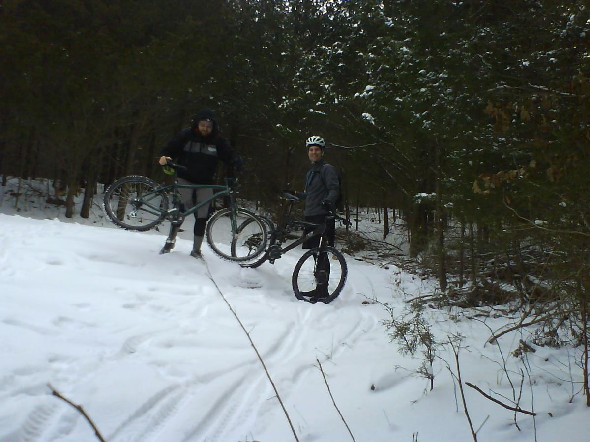 Two cyclists stand on a snowy trail in a forested area. One man is holding a mountain bike while raising it slightly off the ground, and the other is holding a bike with both hands. Snow covers the ground, and trees surround them, creating a winter scene. Sac River Trail mountain bike trail.