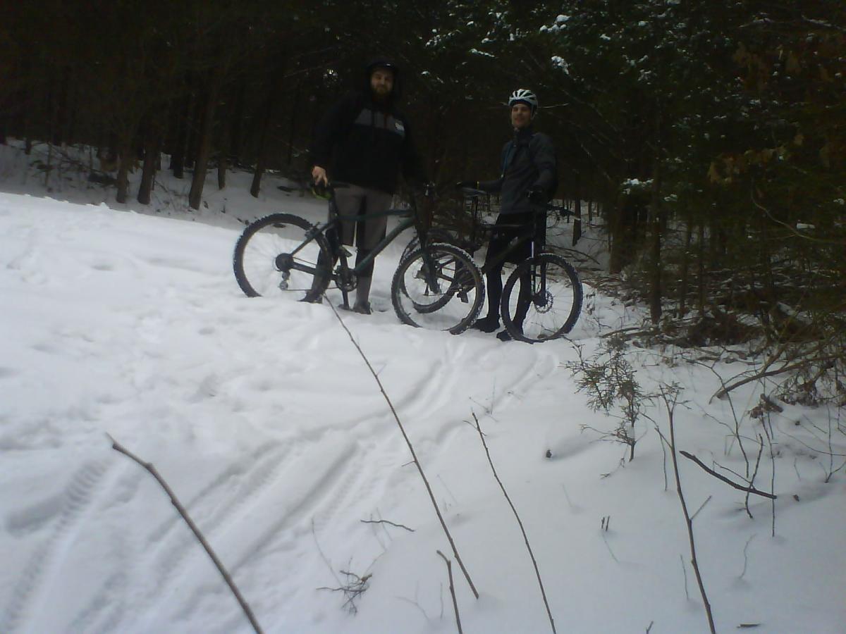 Two individuals stand next to their mountain bikes on a snowy trail surrounded by trees. The ground is covered in fresh snow, and tire tracks can be seen leading through the snow. One person is wearing a dark hoodie, while the other is dressed in a light jacket and a helmet. Sac River Trail mountain bike trail.