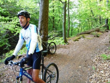 A mountain biker in a cycling outfit stands beside his bike on a forest trail surrounded by green trees. The terrain is gravelly with patches of leaves on the ground. Tower Trail mountain bike trail.