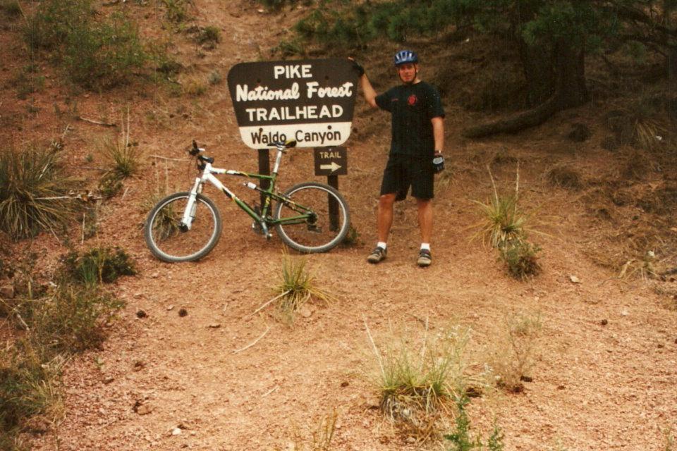 A person in cycling gear stands next to a sign for the Pike National Forest Trailhead, indicating the Waldo Canyon trail. A mountain bike is positioned beside them on a dirt path surrounded by sparse vegetation and trees. Waldo Canyon mountain bike trail.