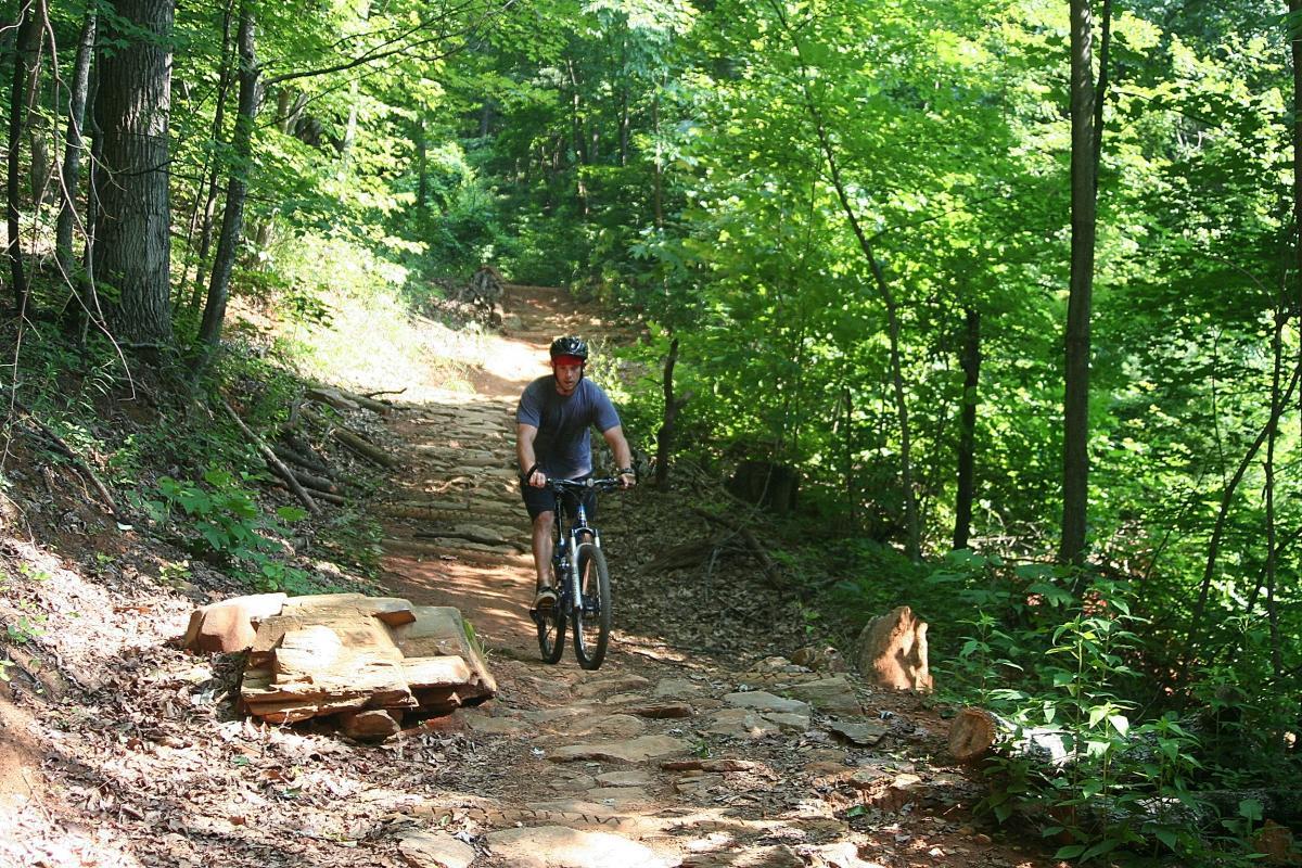 A person riding a mountain bike on a rocky trail surrounded by lush green trees. The trail is uneven with scattered rocks and natural debris, indicating a scenic outdoor environment. Bent Creek mountain bike trail.