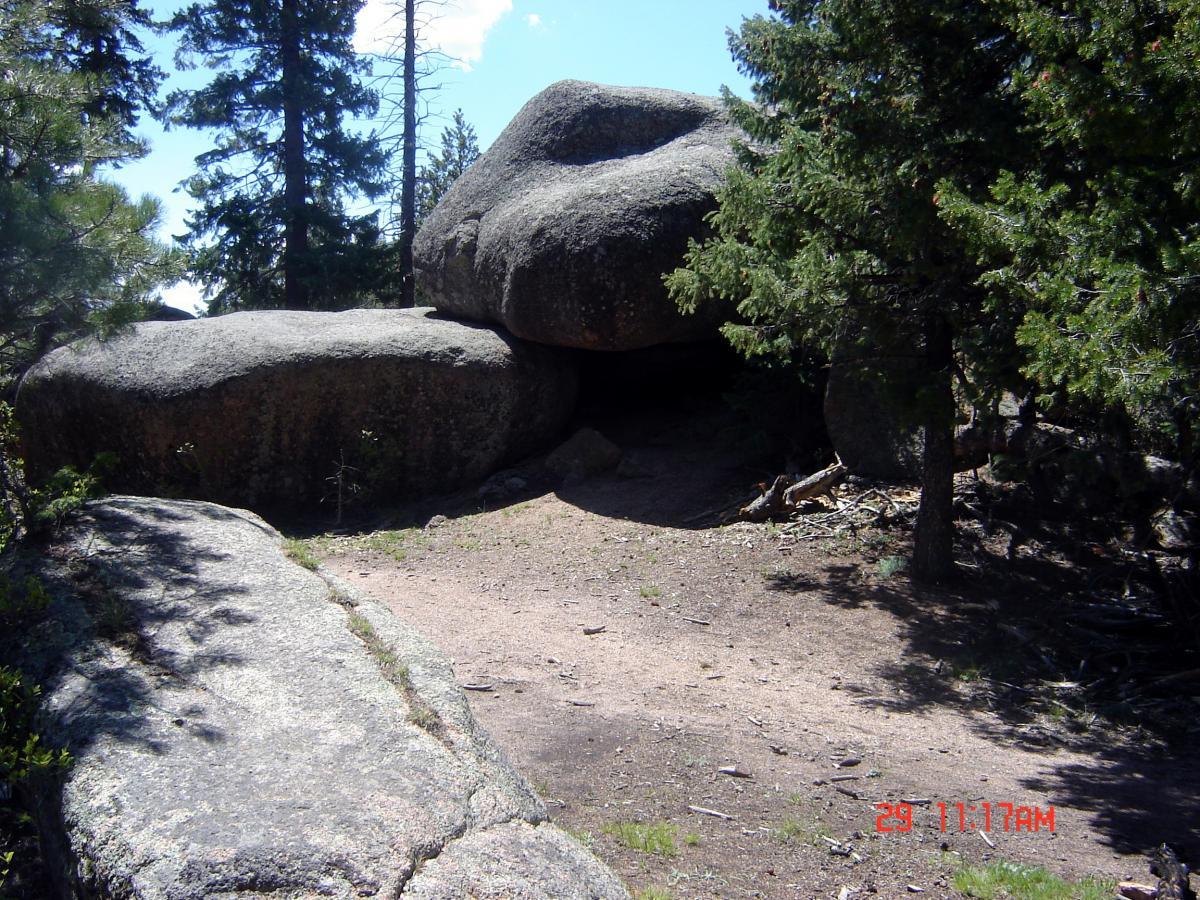 Large granite boulders partially covered by trees, with a clear pathway leading through them. Sunlight filters through the foliage, illuminating the textured surfaces of the rocks and the surrounding earthy ground. Buffalo Creek mountain bike trail.