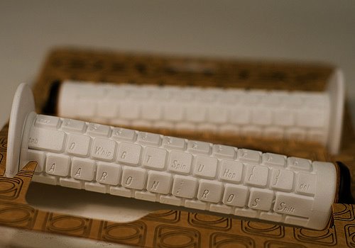 Close-up of two white bicycle handlebar grips designed with raised keyboard-like keys. The grips are placed on a patterned brown surface, highlighting their unique design.