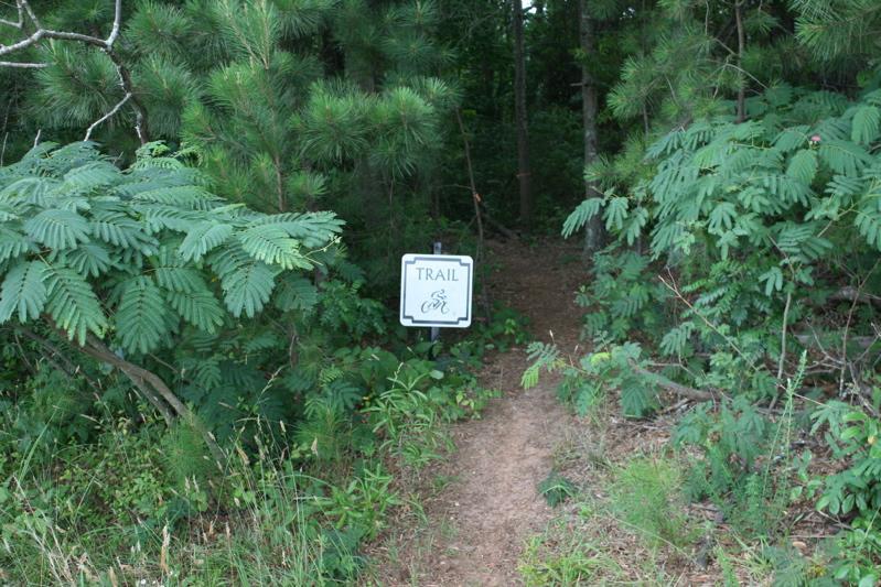 A dirt pathway leading into a wooded area, marked by a sign that reads "TRAIL" with an image of a bicycle. Surrounding vegetation includes various green plants and trees, indicating a natural environment. Kiwanis Fairground mountain bike trail.