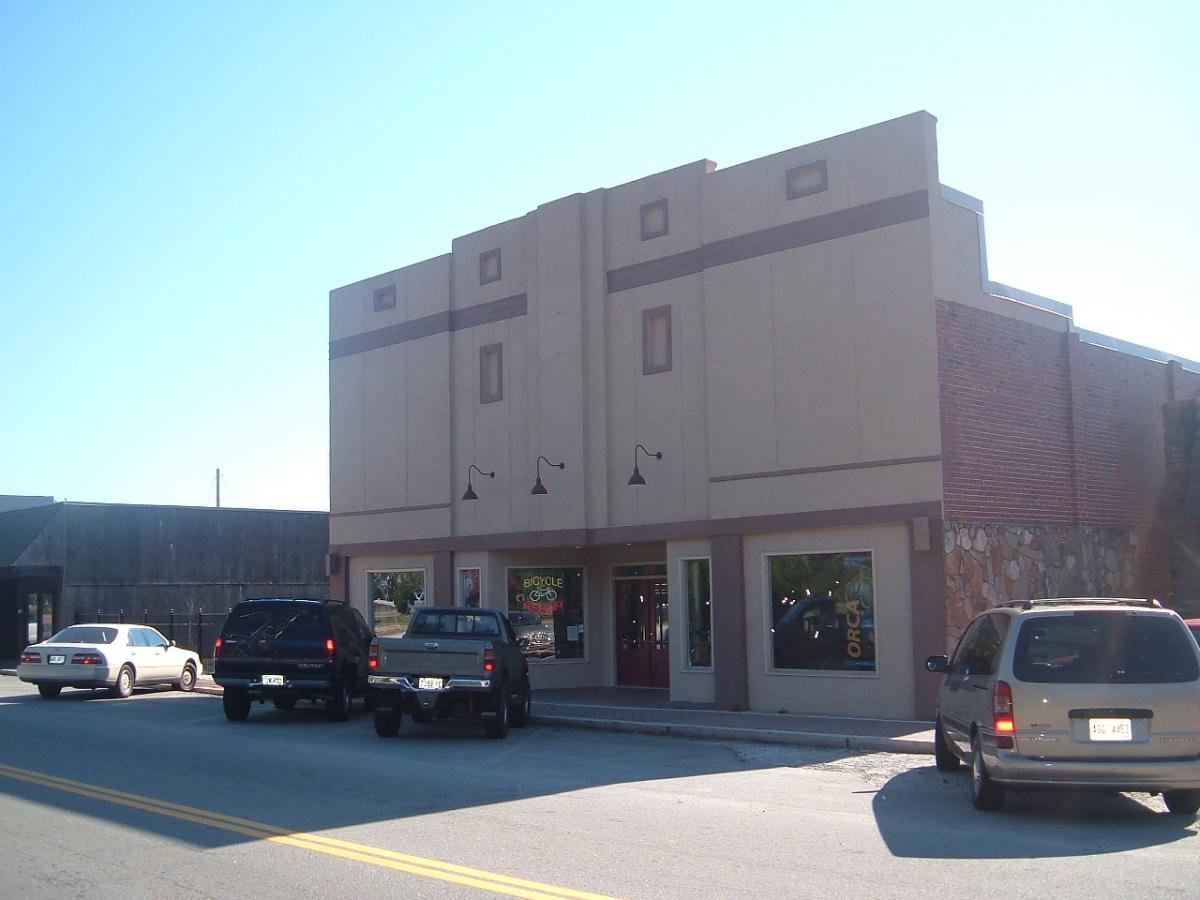 A small building with a light-colored exterior and decorative elements at the top, featuring two large windows framed by brown trim. One window displays a sign for "Bicycle" and another shows signs for "Orca." Several parked vehicles, including black SUVs and a minivan, line the street in front. The sky is clear and blue, creating a bright, sunny atmosphere.
