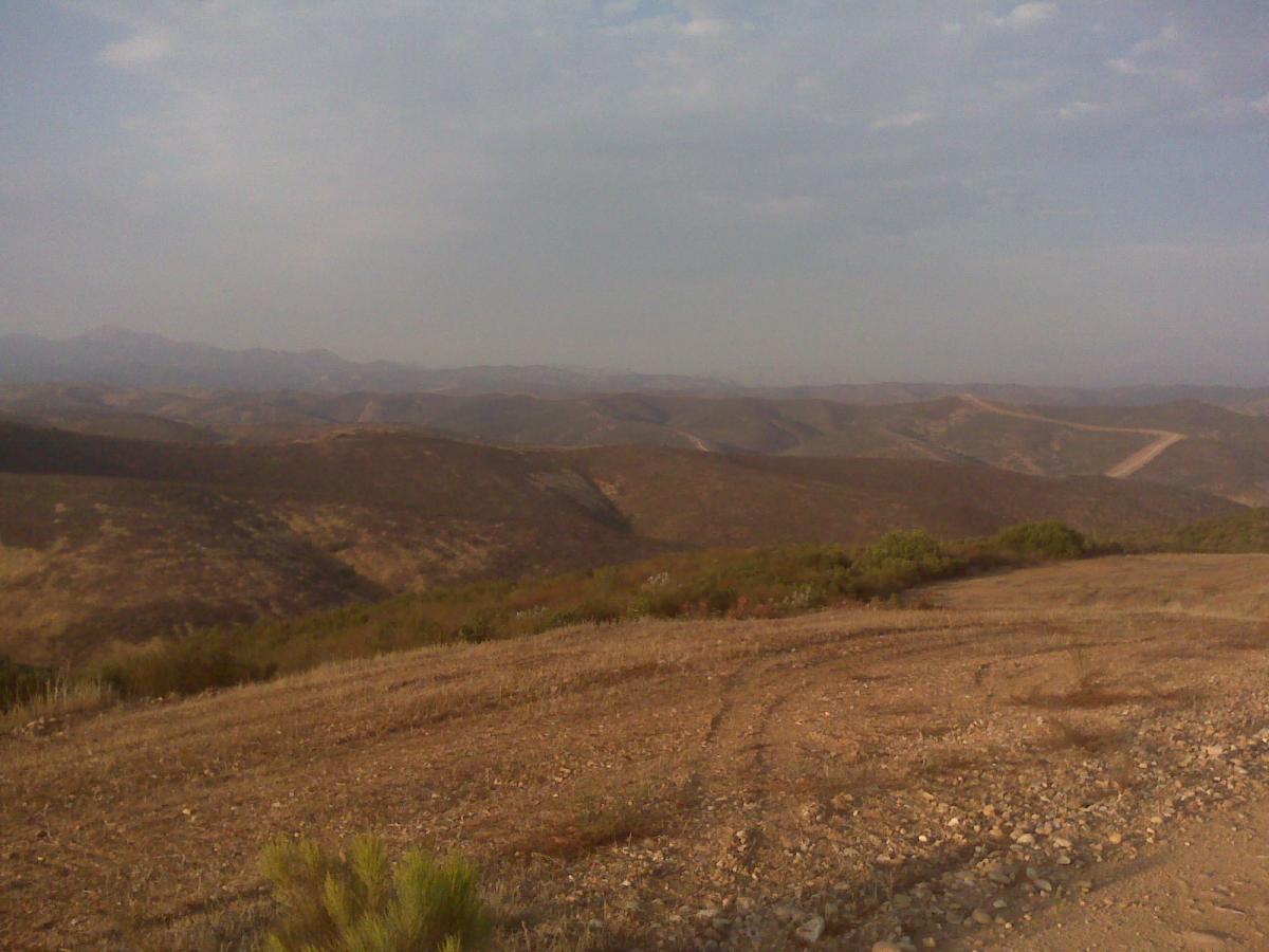 Vast mountainous landscape under a cloudy sky, featuring rolling hills covered in dry grass and sparse vegetation. The foreground shows dirt and gravel with tire tracks, while the background reveals distant mountains fading into the horizon. Spring Canyon mountain bike trail.