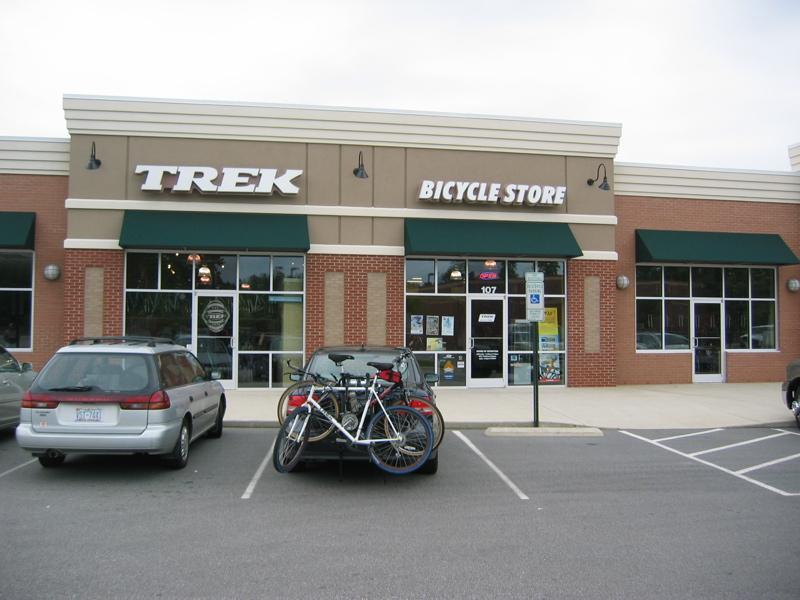 A view of a Trek bicycle store, featuring large signage with the words "TREK" and "BICYCLE STORE." The storefront has green awnings, large windows displaying merchandise, and signs. In the foreground, two parked vehicles, including one with bicycles mounted on a rack, are visible in the parking lot.