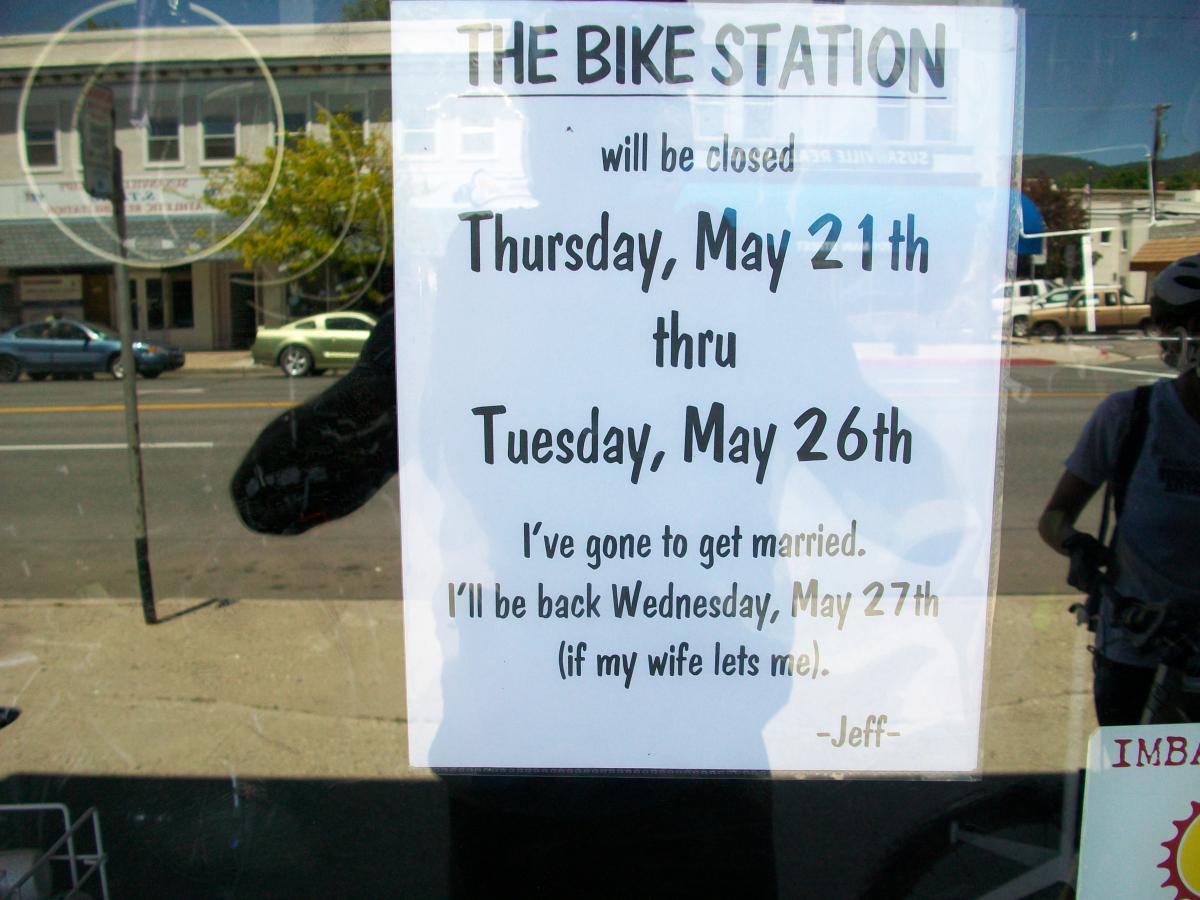 A handwritten sign in a shop window announcing that "The Bike Station" will be closed from Thursday, May 21st through Tuesday, May 26th. The sign humorously states, "I