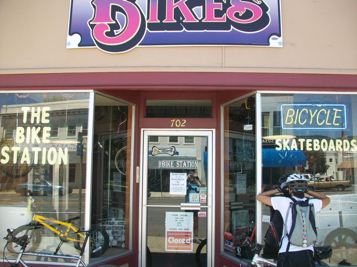 A storefront with signs reading "THE BIKE STATION" and "BICYCLE SKATEBOARDS." A person wearing a helmet and backpack stands in front, preparing their bicycle. The entrance is closed with a notice on the door. The window displays bicycles and accessories.