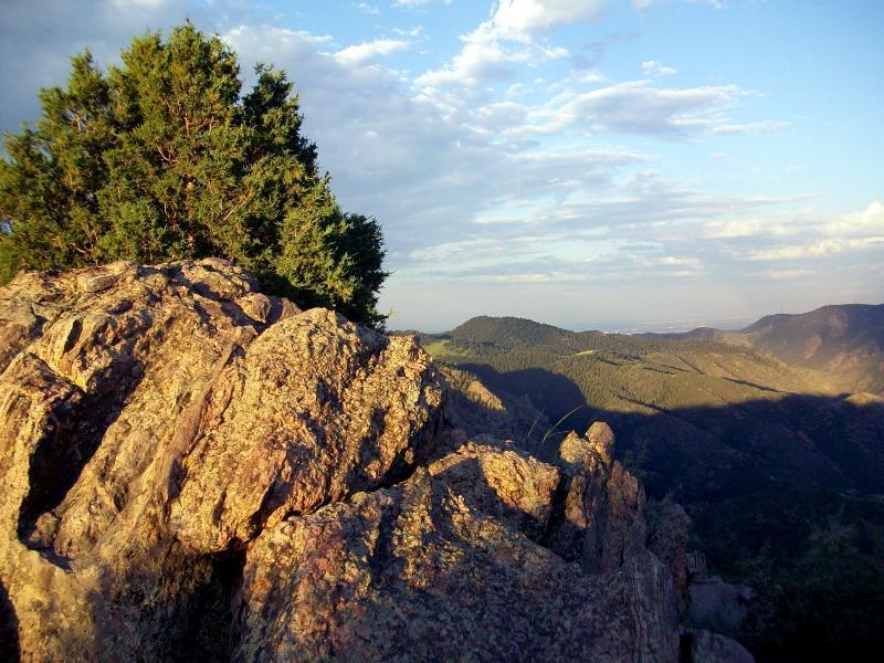 A rocky cliff with a small tree on top, overlooking a vast mountainous landscape under a partly cloudy sky during sunset. Centennial Cone Park mountain bike trail.
