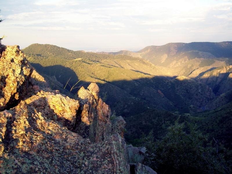 A scenic view of rolling mountains and valleys, with sunlight casting shadows on rocky outcrops in the foreground. The landscape is filled with green trees and hills, stretching into the distance under a partly cloudy sky. Centennial Cone Park mountain bike trail.