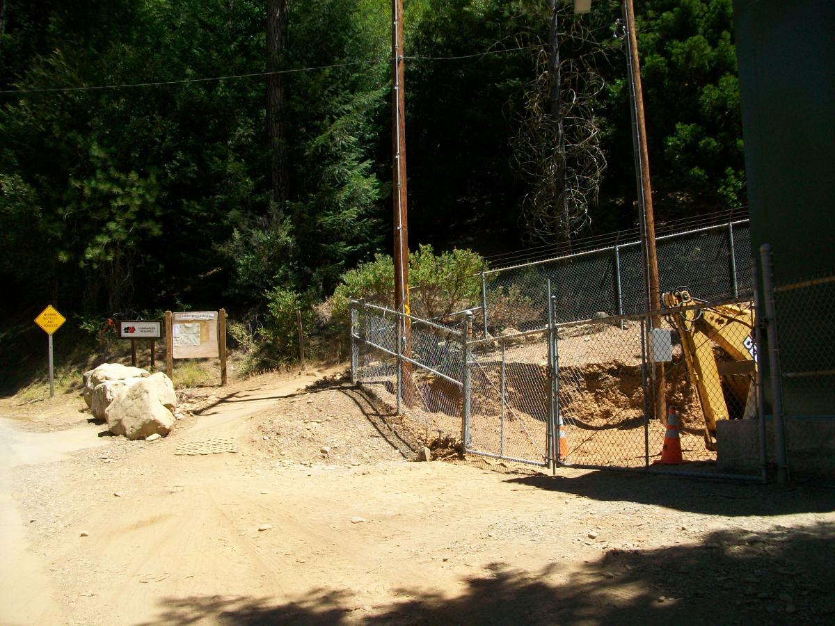A dirt path leading to a fenced construction area surrounded by trees. A sign with a map and warnings is visible on the left, and large rocks are placed at the entrance. A piece of construction equipment is partially visible behind the fence. 1st / 2nd / 3rd Divide mountain bike trail.