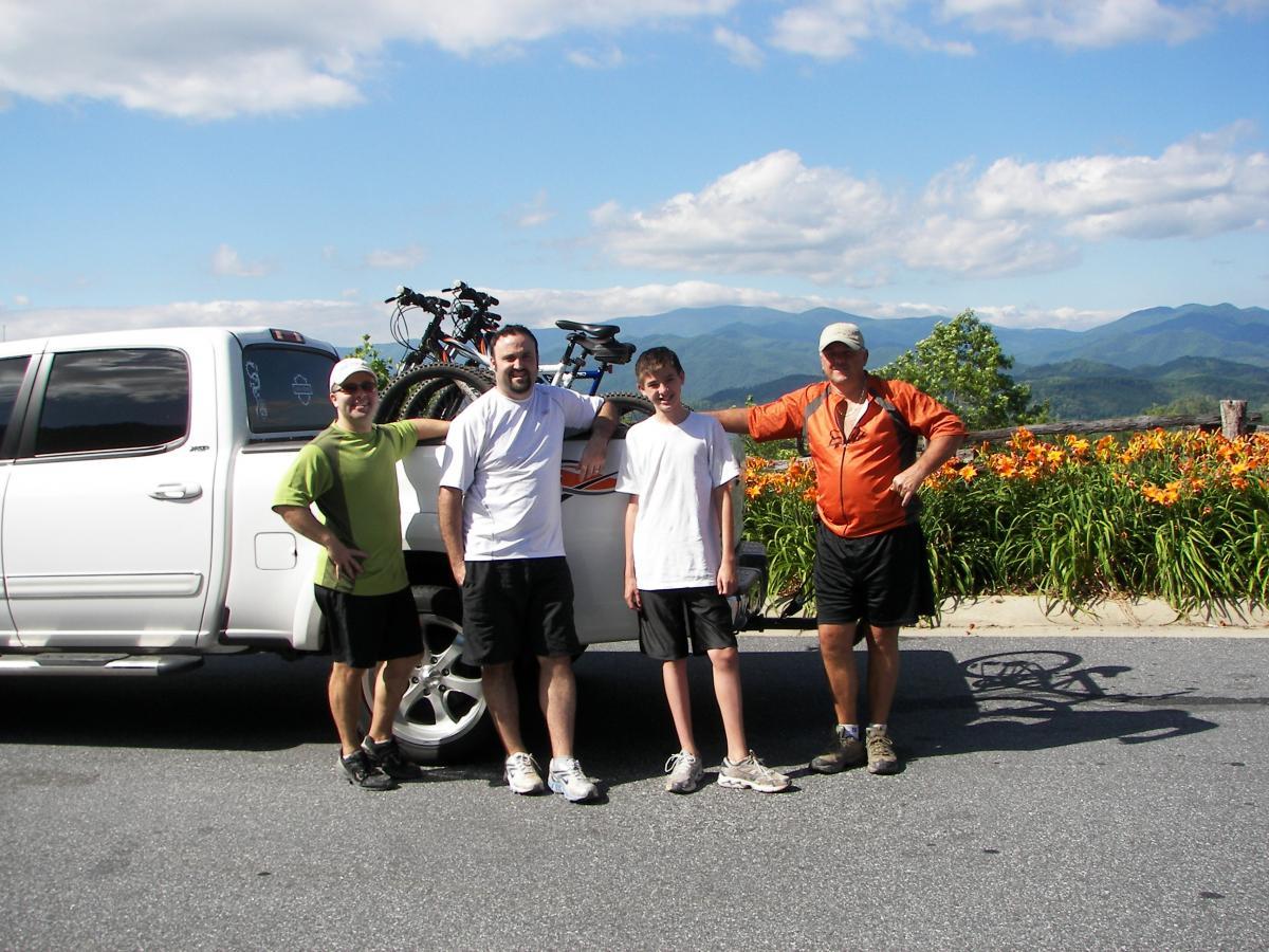 A group of four people standing next to a white pickup truck with mountain scenery in the background. The individuals are smiling and dressed in casual athletic clothing, with bicycles visible in the truck bed. Bright flowers line the roadside under a clear blue sky. Tsali Right Loop mountain bike trail.