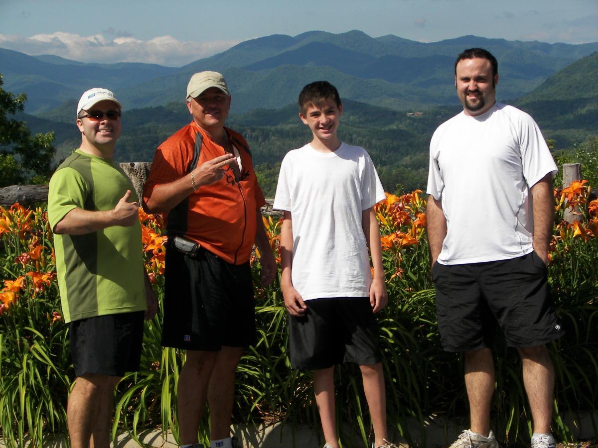 Four individuals stand together outdoors, smiling in front of a vibrant display of orange flowers. They are positioned against a backdrop of rolling mountains and a clear blue sky. The group consists of two men, one of whom is wearing an orange shirt, a teenage boy in a white t-shirt, and another man in a white shirt. The scene suggests a joyful moment, likely taken during a family outing or hike. Tsali Right Loop mountain bike trail.